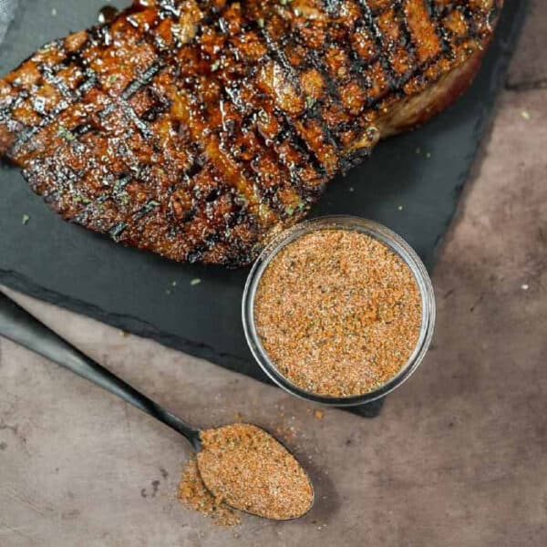 Grilled steak with perfect crisscross grill marks sits on a dark slate, showcasing the mouthwatering essence of Texas Roadhouse Steak Seasoning. A small container of this savory blend is beside it, with a spoonful waiting nearby on the textured gray surface.