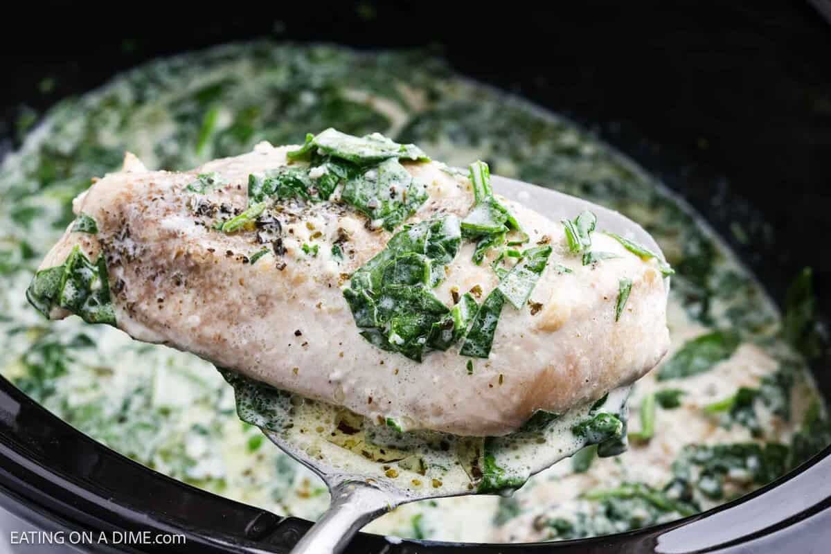 A spoon holds a creamy, cooked chicken breast topped with chopped spinach and herbs above a slow cooker filled with a rich spinach and cream sauceāperfect for this Crock Pot creamy parmesan chicken recipe.