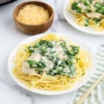 A white plate of spaghetti topped with creamy spinach and tender crock pot creamy parmesan chicken, with a bowl of shredded Parmesan cheese in the background and a green-striped napkin beside the plate.