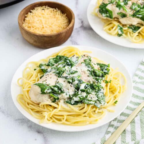 A white plate of spaghetti topped with creamy spinach and tender crock pot creamy parmesan chicken, with a bowl of shredded Parmesan cheese in the background and a green-striped napkin beside the plate.