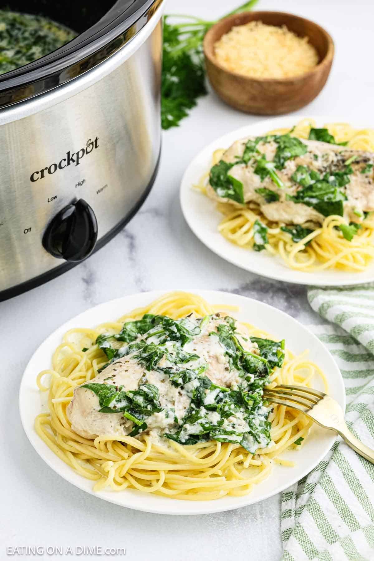 A plate of spaghetti topped with Crock Pot creamy parmesan chicken sits in front of a slow cooker. Another similar plate and a bowl of grated cheese are in the background, along with a striped napkin and fresh parsley.