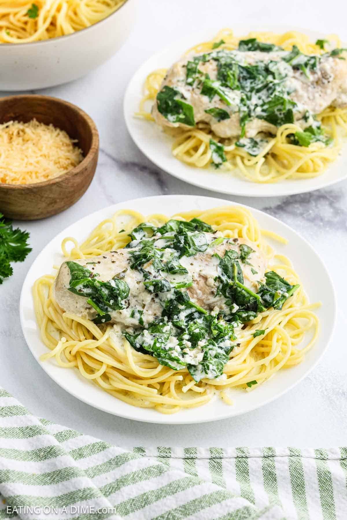 Two plates of spaghetti topped with creamy spinach and chicken, made from a crock pot creamy parmesan chicken recipe, sit next to a small wooden bowl of grated cheese on a white surface with a green striped towel in the corner.