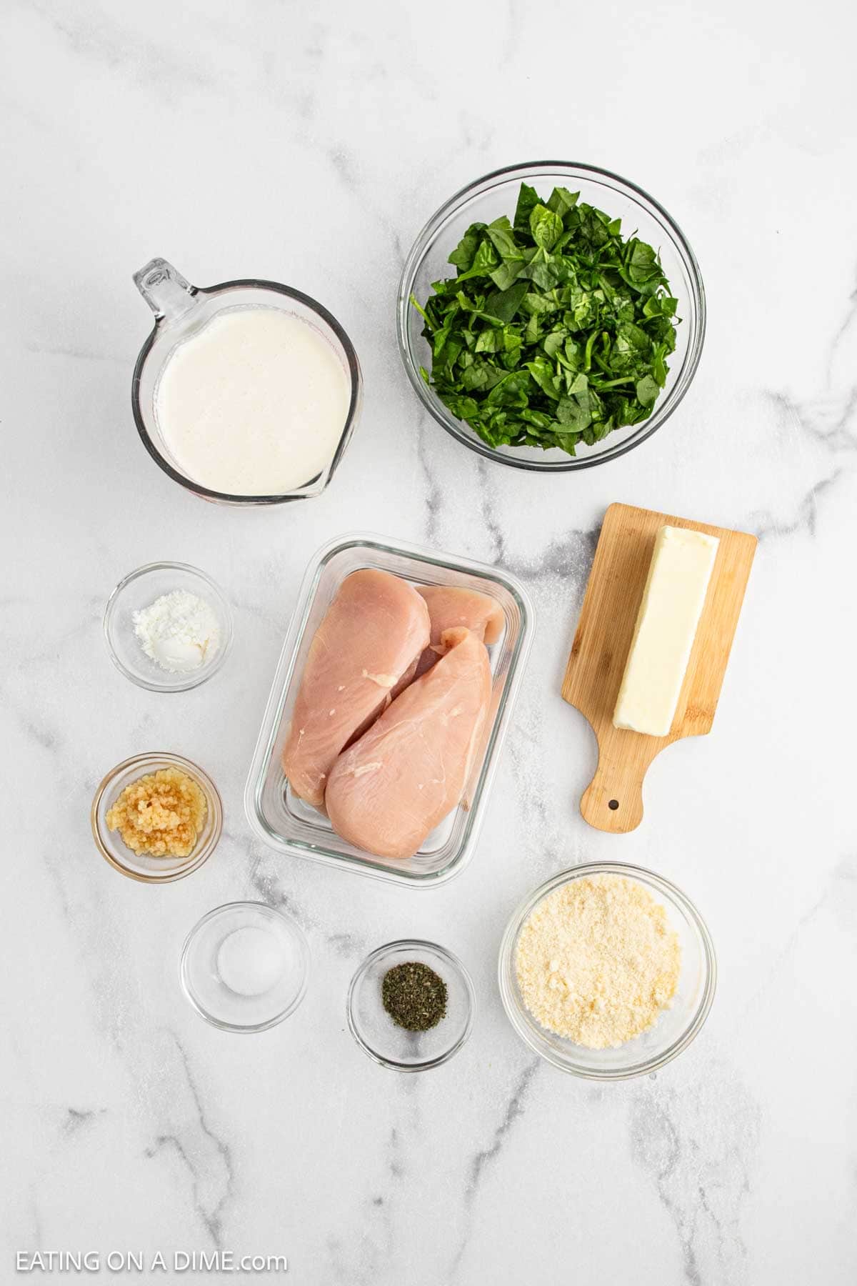 Top-down view of ingredients for a Crock Pot creamy Parmesan chicken recipe: raw chicken breasts, chopped spinach, grated Parmesan cheese, a stick of butter, minced garlic, heavy cream, black pepper, and cornstarch in a small bowl.