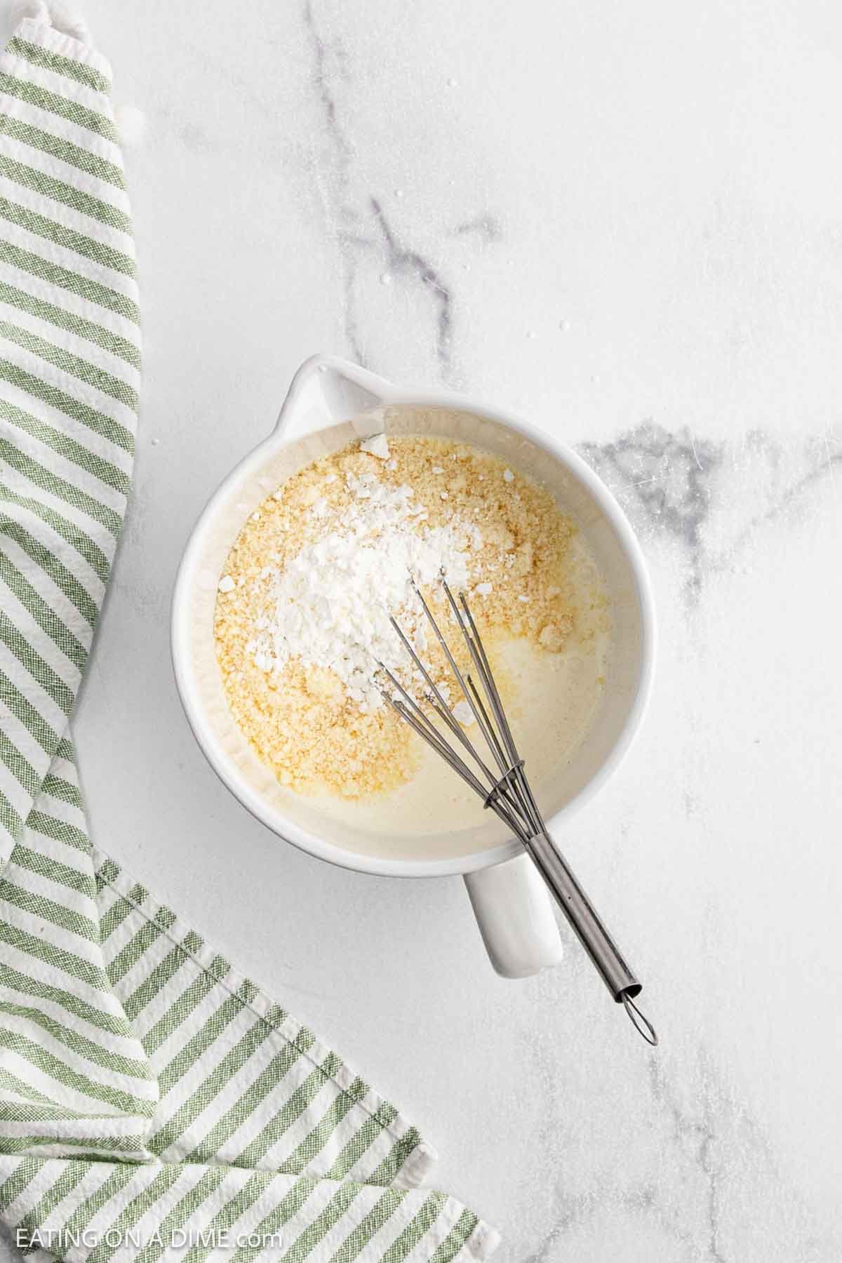 A white bowl with flour, cornmeal, and cream mixture being whisked for a Crock Pot creamy Parmesan chicken recipe, next to a green and white striped kitchen towel on a marble surface.