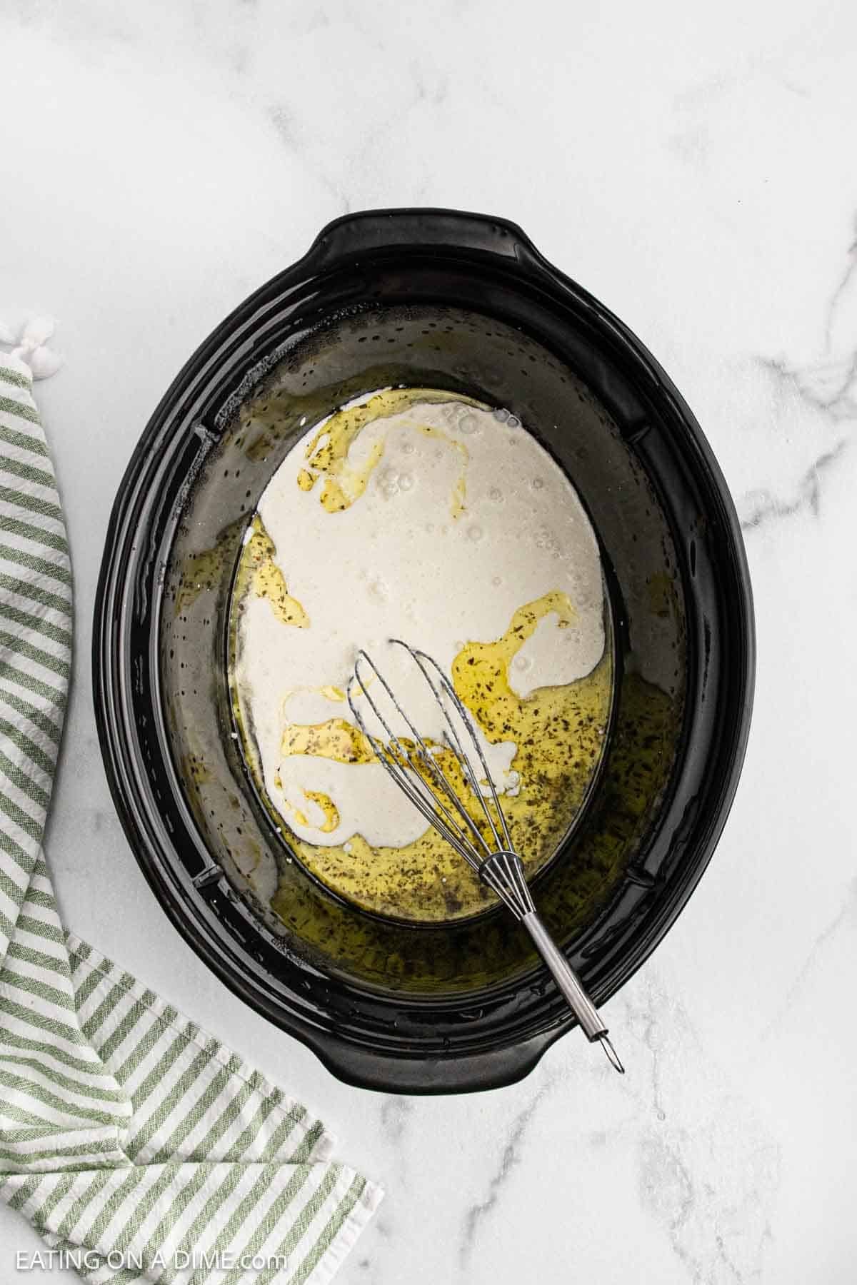 A black slow cooker filled with creamy liquid, oil, and seasonings for a crock pot creamy parmesan chicken recipe, with a metal whisk inside. A green and white striped towel rests beside the cooker on a white marble surface.
