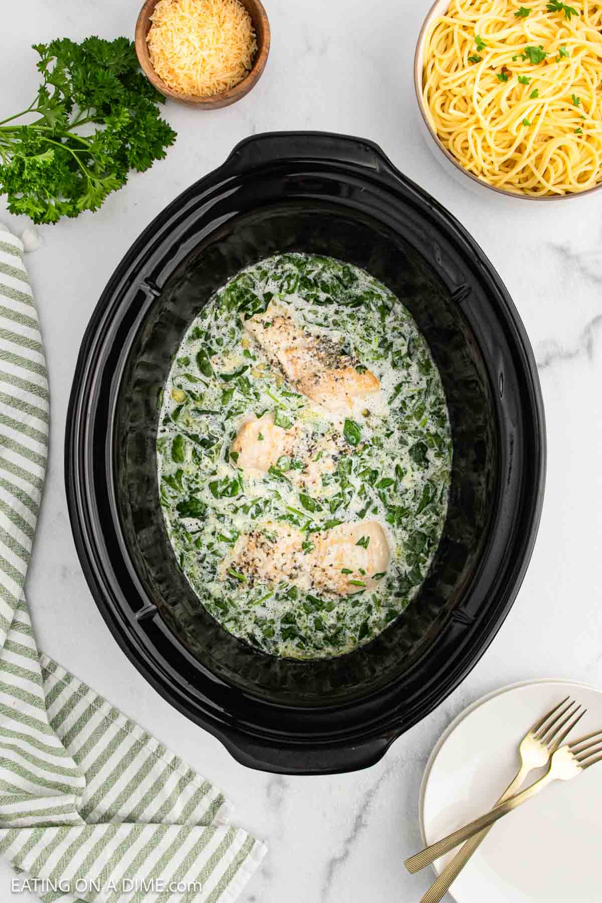 A black slow cooker showcases this Crock Pot creamy parmesan chicken recipe with spinach, surrounded by shredded cheese, cooked spaghetti, parsley, a striped towel, and a fork and knife on a plate.
