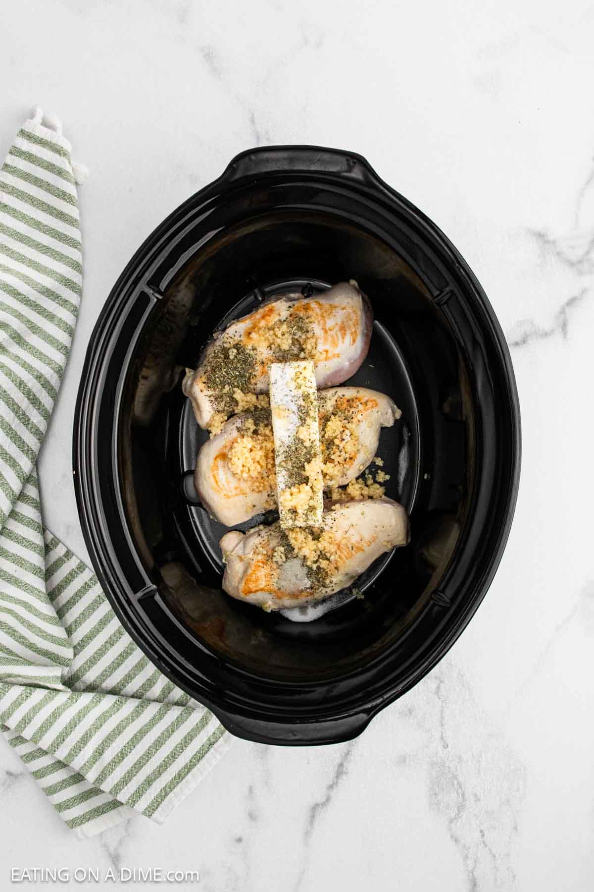 Three raw chicken breasts topped with seasonings and a block of butter sit in a black slow cooker on a white marble surfaceāperfect for your next Crock pot creamy parmesan chicken recipe. A green and white striped cloth is positioned to the left.