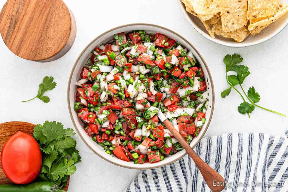 A bowl of easy pico de gallo made with diced tomatoes, onions, cilantro, and jalapeños. A wooden spoon rests in the bowl. Surrounding it are tortilla chips, a whole tomato, jalapeño, cilantro, and a striped cloth.