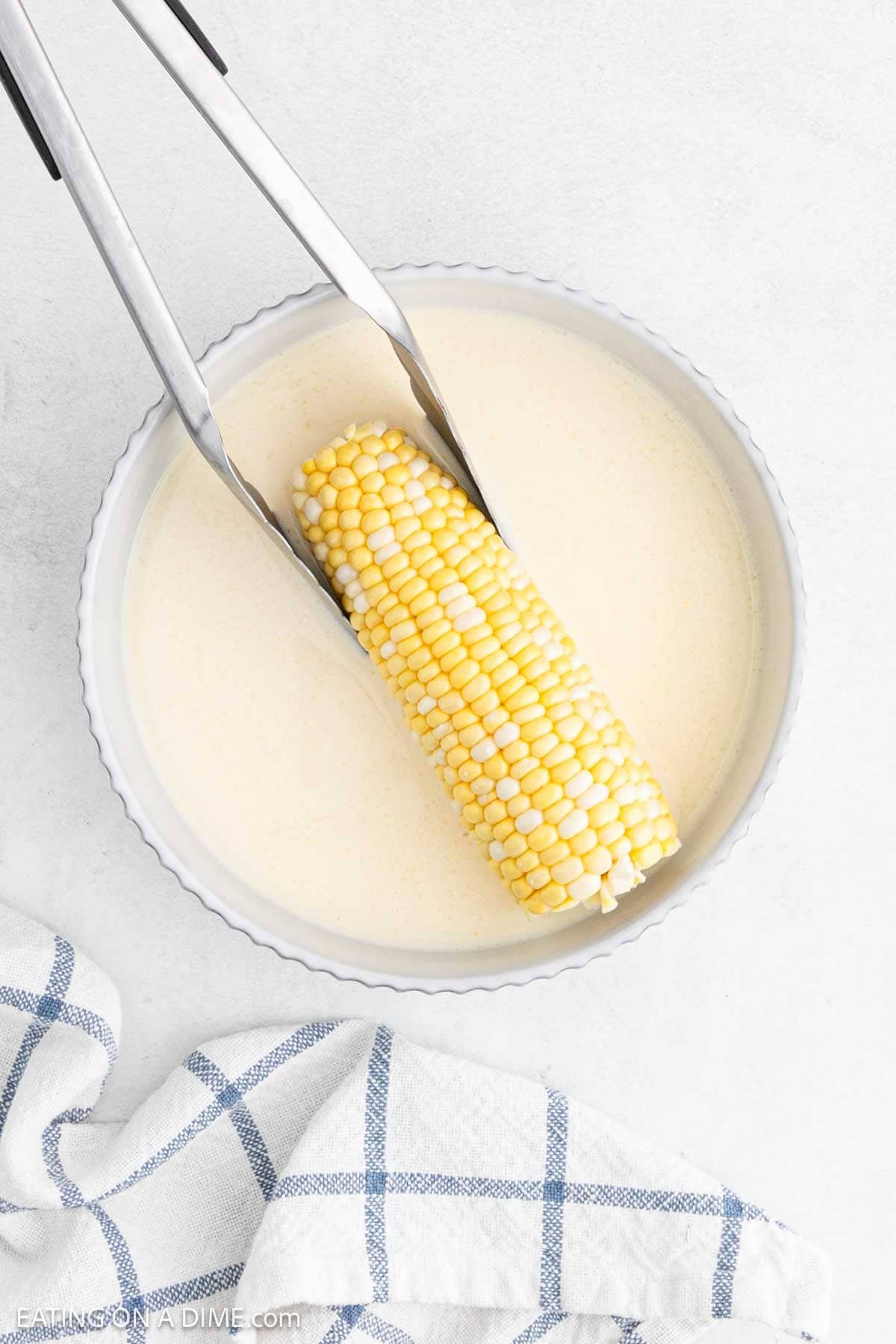 Corn on the cob with tongs holding on to it being dipped into the milk mixture