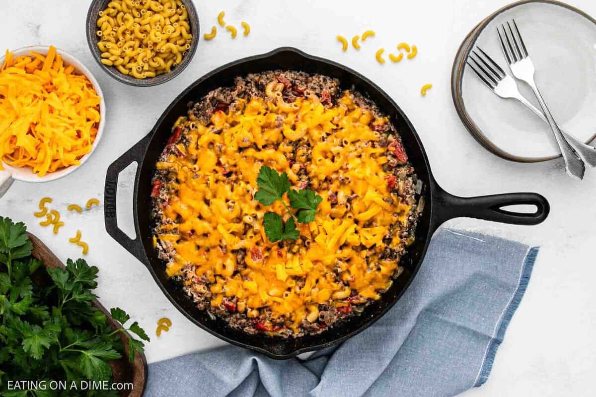 Sausage mac and cheese in a cast iron skillet topped with melted cheese with small bowls of shredded cheese and macaroni on the side.