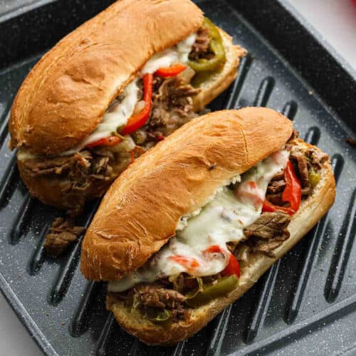 Two crock pot Philly cheesesteak sandwiches on a grill pan, filled with tender sliced beef, melted cheese, red and green bell peppers inside toasted hoagie rolls.
