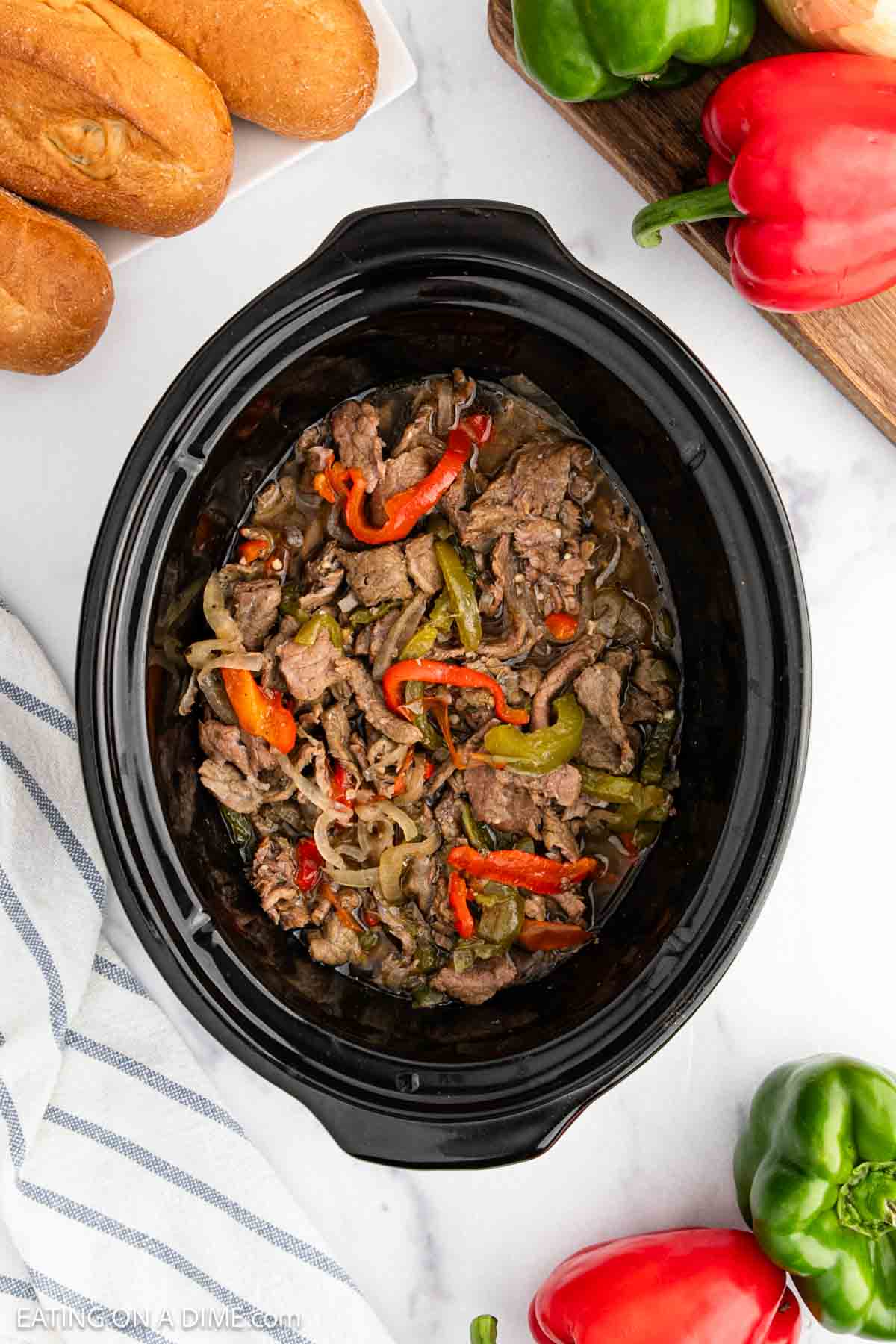 A slow cooker filled with cooked beef strips, red and green bell peppers, and onions for a delicious crock pot Philly cheesesteak sandwich, surrounded by sandwich rolls and fresh bell peppers on a white countertop.