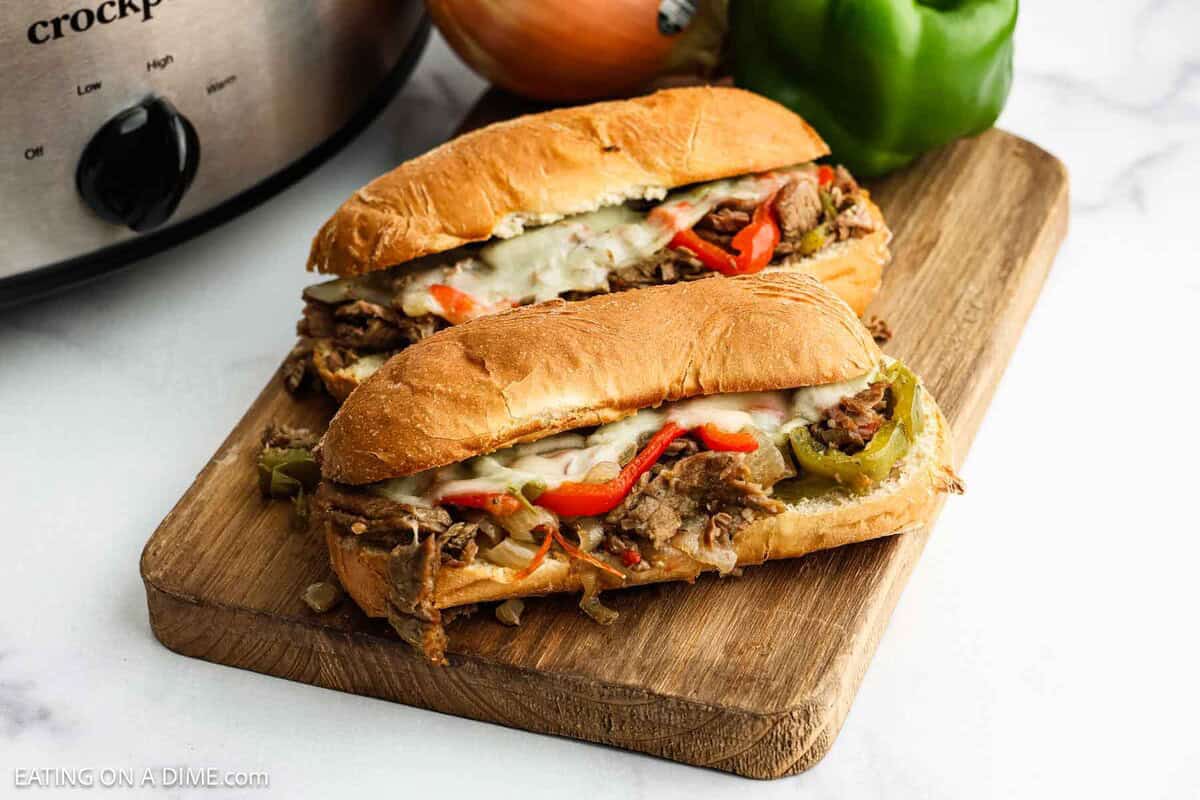 Two hoagie sandwiches filled with shredded beef, melted cheese, and bell peppers sit on a wooden board—a classic crock pot Philly cheesesteak sandwich. An onion, green bell pepper, and slow cooker are visible in the background.