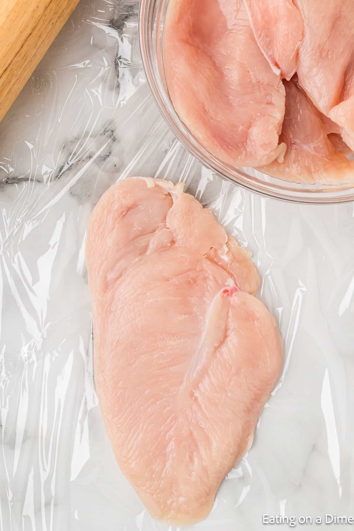 Chicken breast on plastic wrap with a bowl of chicken on the side