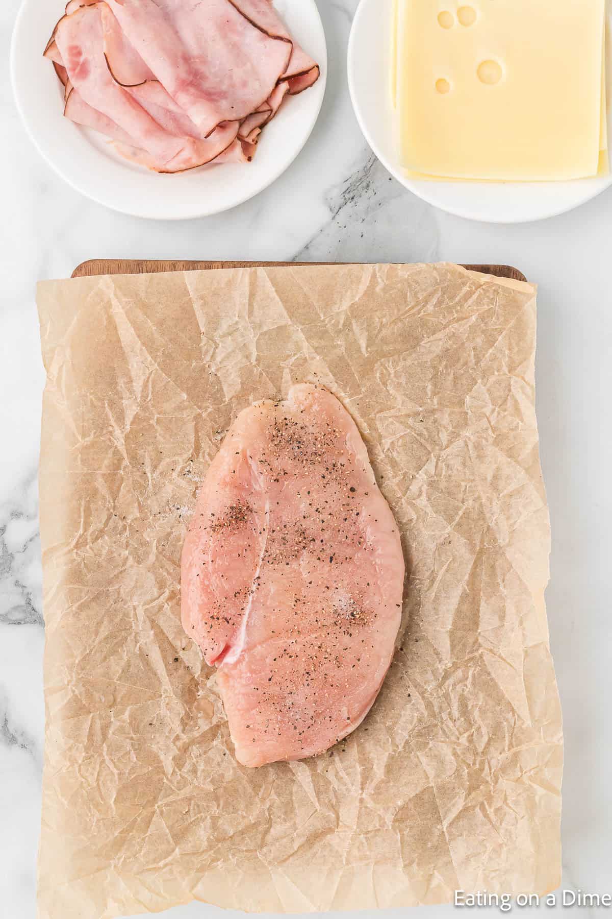 Chicken breast on a piece of parchment paper seasoned with a plate of deli ham and slices of cheese on the side