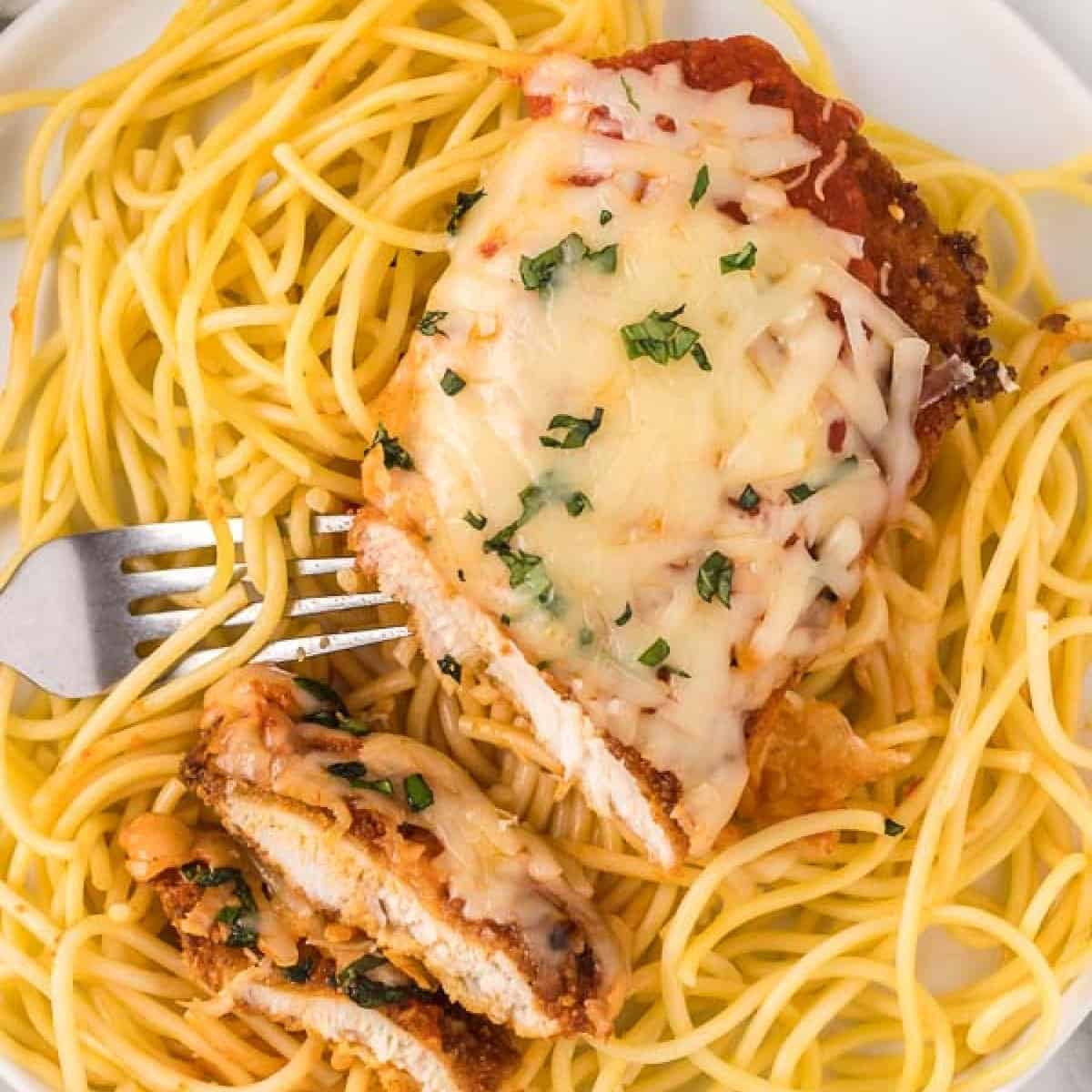 A plate of spaghetti topped with a slice of chicken parmesan, covered in melted cheese and garnished with herbs. A fork holds a portion of the delicious Chicken Parmesan recipe, and the dish is served on a white plate.