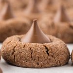 A close-up of Chocolate Kiss Cookies showcases a sugar-coated, cracked surface and a chocolate kiss in the center. More cookies are blurred in the background.