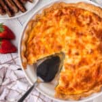 A baked quiche made from a delicious quiche recipe sits on a white plate with a slice missing. A pie server rests in the empty space, while fresh strawberries, sausages, and a white towel are nearby on the table.