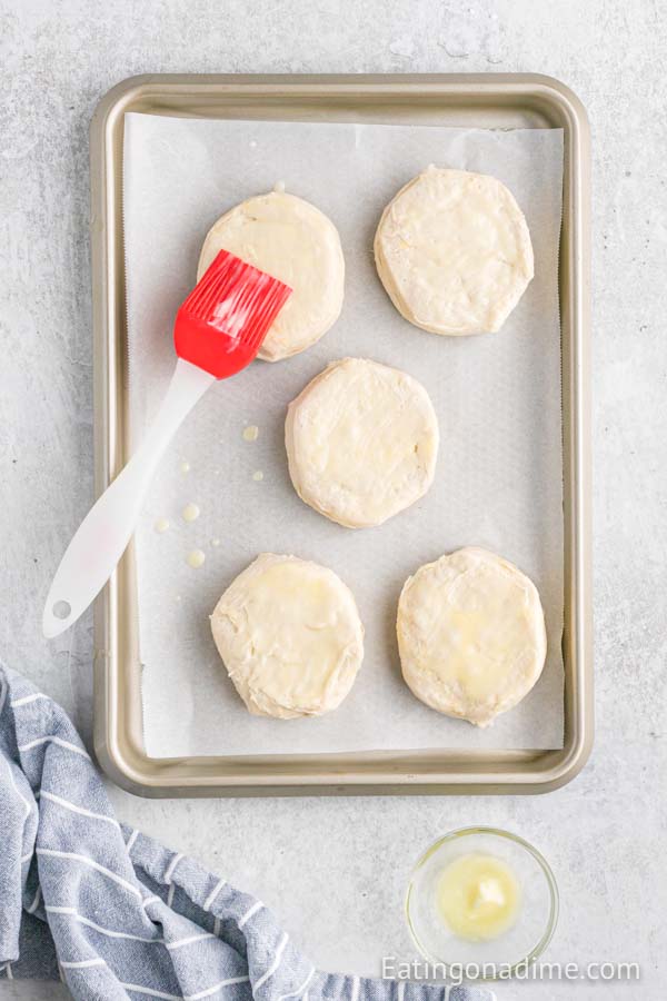 Canned biscuits on a baking sheet being brushed with butter. 