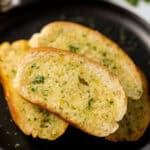 Slices of golden-brown air fryer garlic bread topped with chopped herbs are arranged on a black plate. The bread appears crispy on the edges with a soft, buttery center.