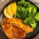 A black plate with two pieces of seasoned, cooked air fryer tilapia, fresh broccoli florets, and three lemon wedges.