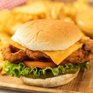 Close-up of a Chick-fil-a Deluxe Chicken Sandwich with cheese, lettuce, tomato, and pickles nestled in a bun. It rests on a wooden surface with crinkle-cut fries in the background and a side of dipping sauce.