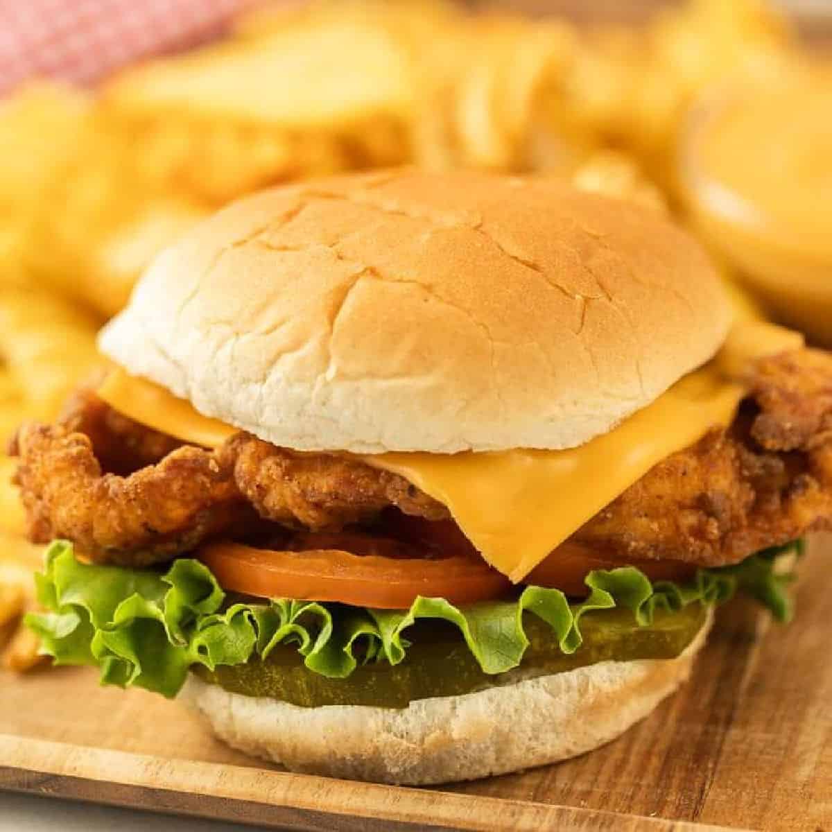 Close-up of a Chick-fil-a Deluxe Chicken Sandwich on a wooden board. The sandwich features a golden-brown fried chicken fillet, cheddar cheese, lettuce, tomato slices, and pickles inside a soft bun. Crinkle-cut fries and dipping sauce sit blurred in the background.
