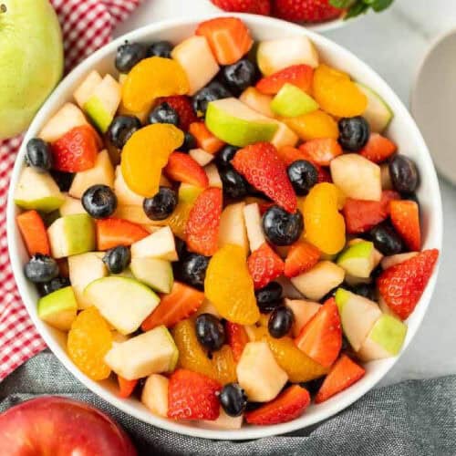 A vibrant fruit salad with strawberries, blueberries, mandarins, apples, and watermelon, served beside fresh strawberries on a red checkered cloth.