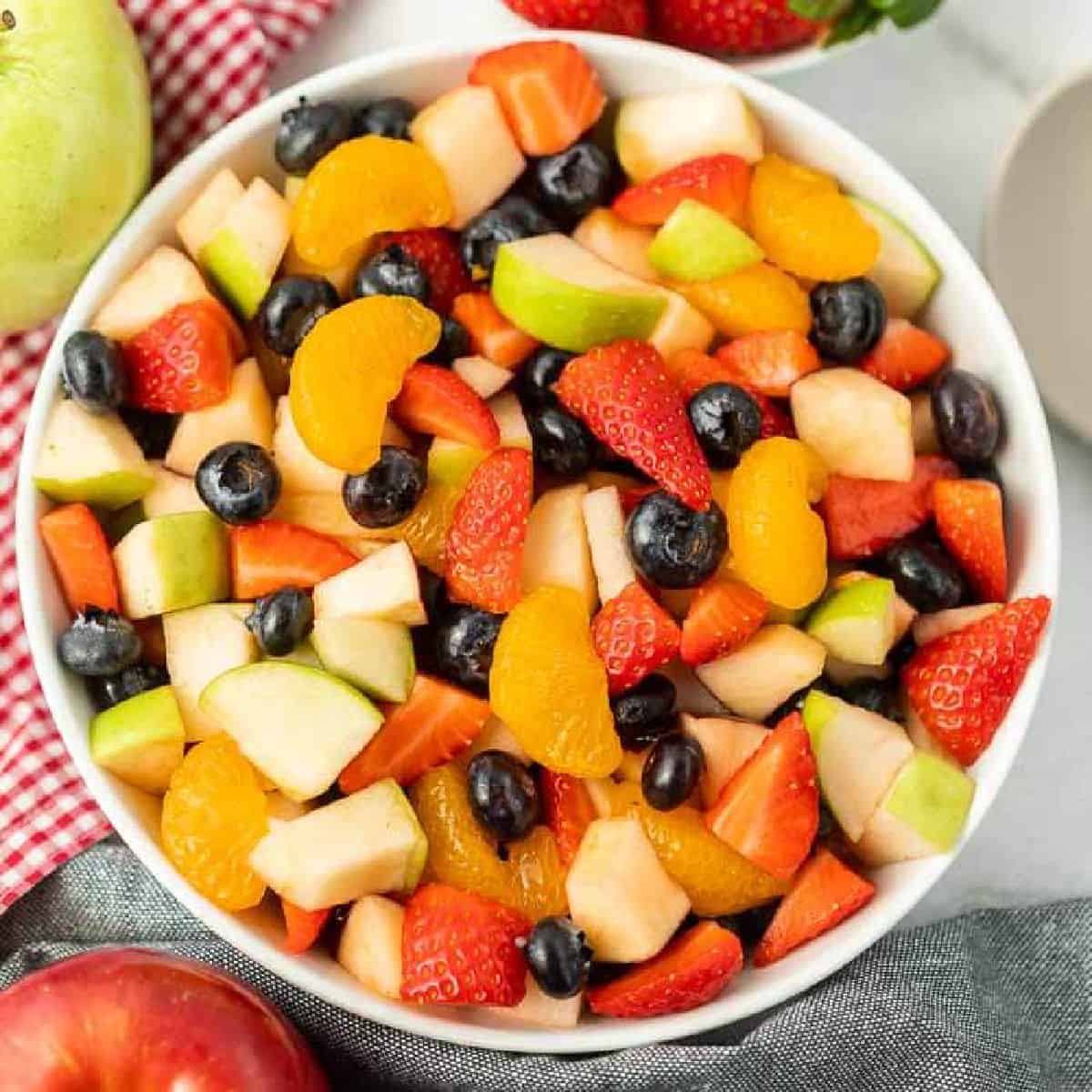 A vibrant fruit salad with strawberries, blueberries, mandarins, apples, and watermelon, served beside fresh strawberries on a red checkered cloth.