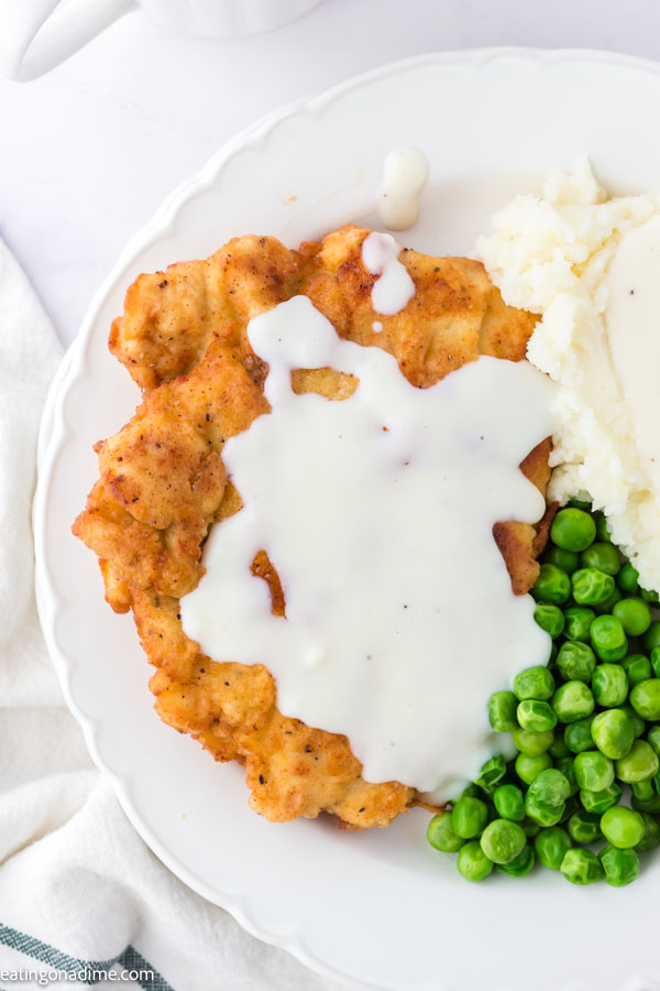 Close up image of chicken fried chicken on a white plate with peas and mashed potatoes and gravy. 