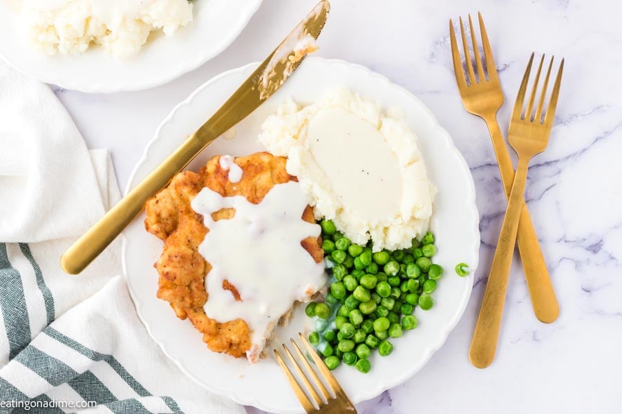 Close up image of chicken fried chicken on a white plate with peas and mashed potatoes and gravy. 