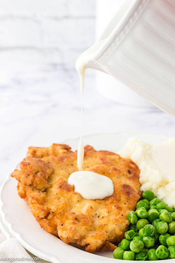 Close up image of chicken fried chicken on a white plate with peas and mashed potatoes and gravy. 
