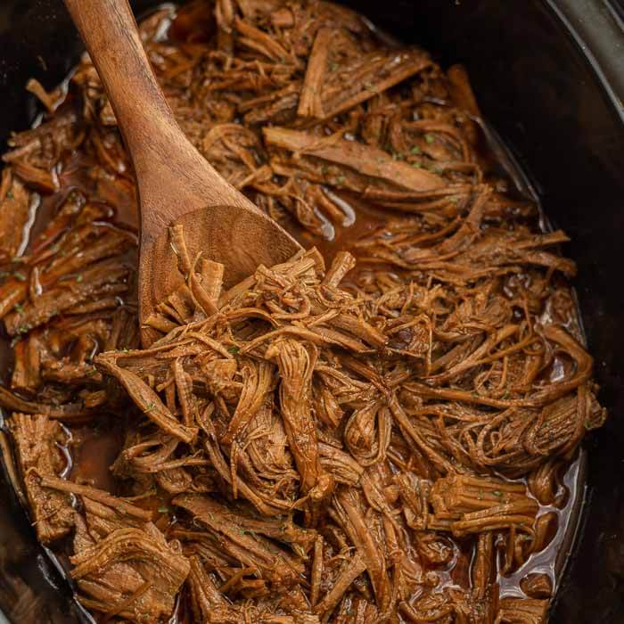 Close up image of shredded beef brisket in a crock pot with a wooden spoon. 