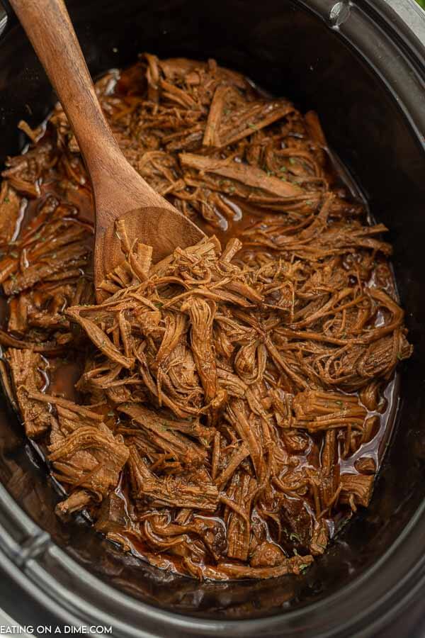 Close up image of shredded beef brisket in a crock pot with a wooden spoon. 