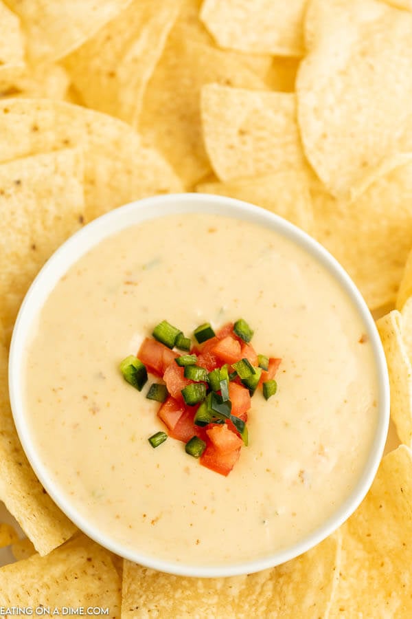 Close up image of queso in a bowl with a side of chips topped with tomatoes and chopped jalapeno