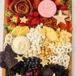 A colorful Red White and Blue Charcuterie Board featuring assorted cheeses, star-shaped cheese slices, strawberries, raspberries, blueberries, nuts, crackers, chocolate-covered pretzels, blue corn chips, marshmallows, and two dips.