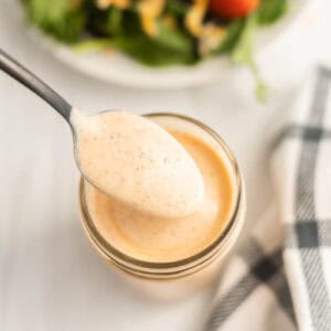 A spoon holding creamy, orange-tinted Southwest Salad Dressing hovers above a glass jar filled with the same dressing; a plate of salad and a checkered cloth are visible in the background.
