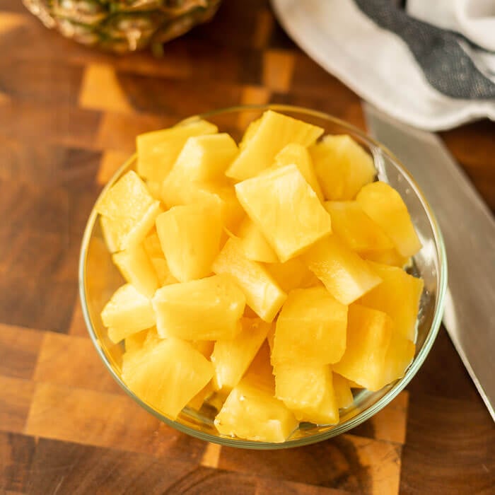 Close up image of a bowl of pineapple chunks on a cutting board with a knife and a whole pineapple