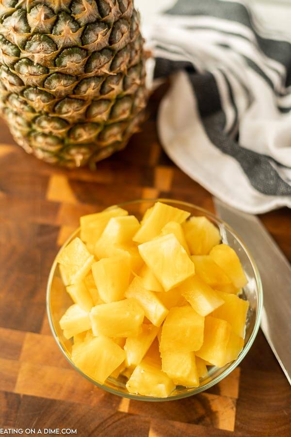 Close up image of a bowl of pineapple chunks on a cutting board with a knife and a whole pineapple