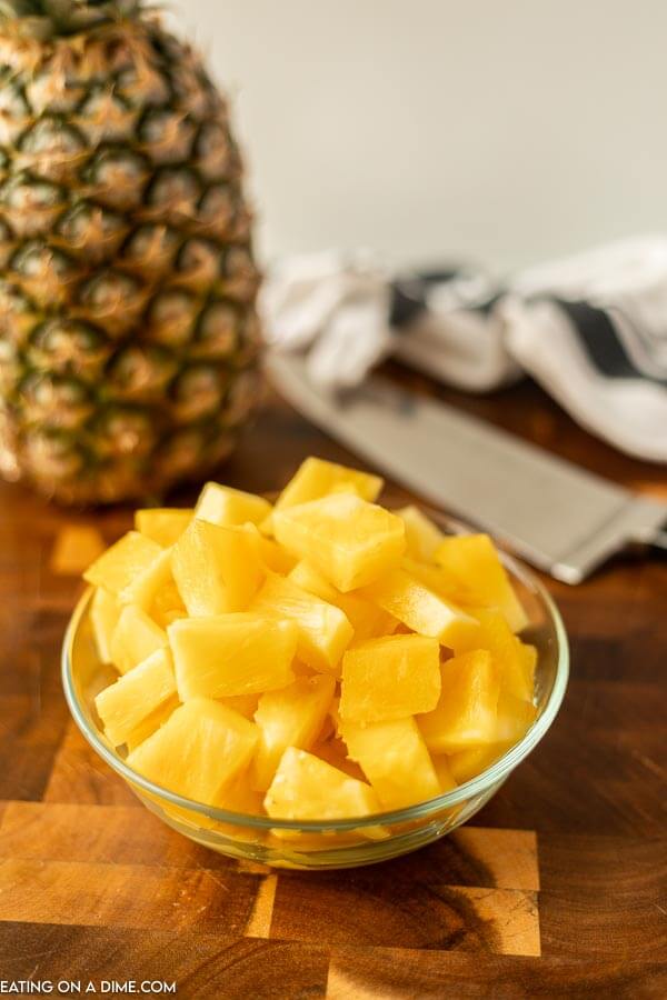 Close up image of a bowl of pineapple chunks on a cutting board with a knife and a whole pineapple