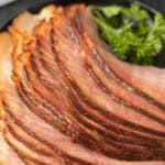 Slices of glazed ham arranged in a circular pattern on a dark plate, garnished with a sprig of fresh parsley. The ham's caramelized crust highlights its texture and color, making you wonder—not just how delicious it looks, but can you freeze ham for later enjoyment?.
