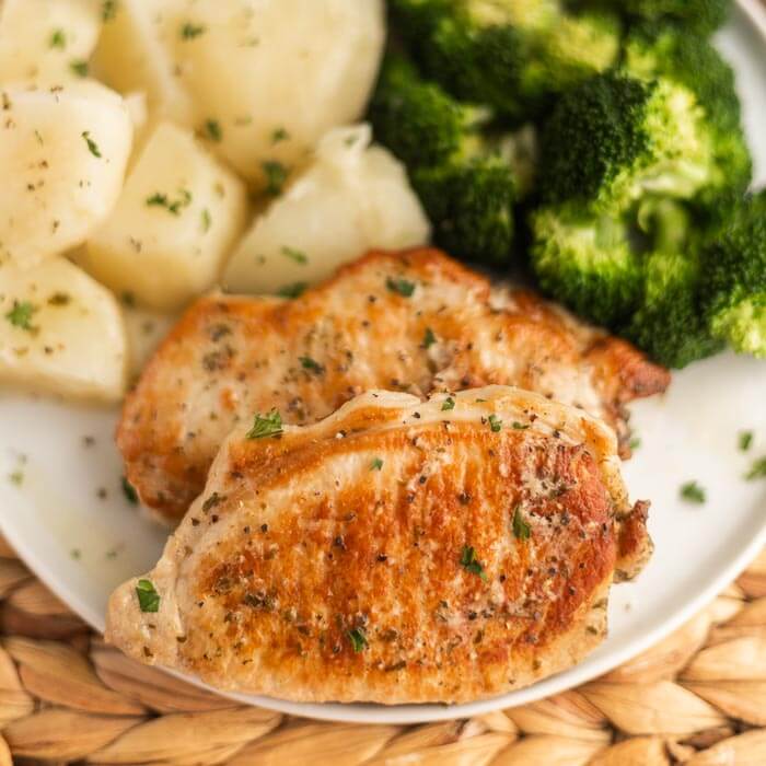 Close up image of pork chops on a white plate with a side of potatoes and broccoli. 