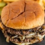 A close-up of a double cheeseburger on a soft bun, featuring juicy beef patties and grilled onions. Pickles and French fries are slightly blurred in the background.