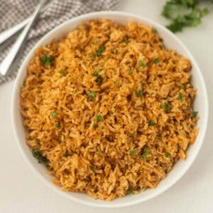 A bowl of fluffy, golden-brown Texas Roadhouse seasoned rice garnished with chopped parsley sits on a light surface, accompanied by a checkered cloth and two silver forks nearby.