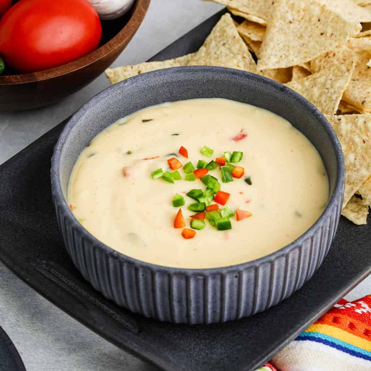 A bowl of creamy Copycat Chipotle Queso Blanco dip topped with chopped red and green peppers, served with tortilla chips on a dark tray. Tomatoes and a bowl of vegetables are in the background.