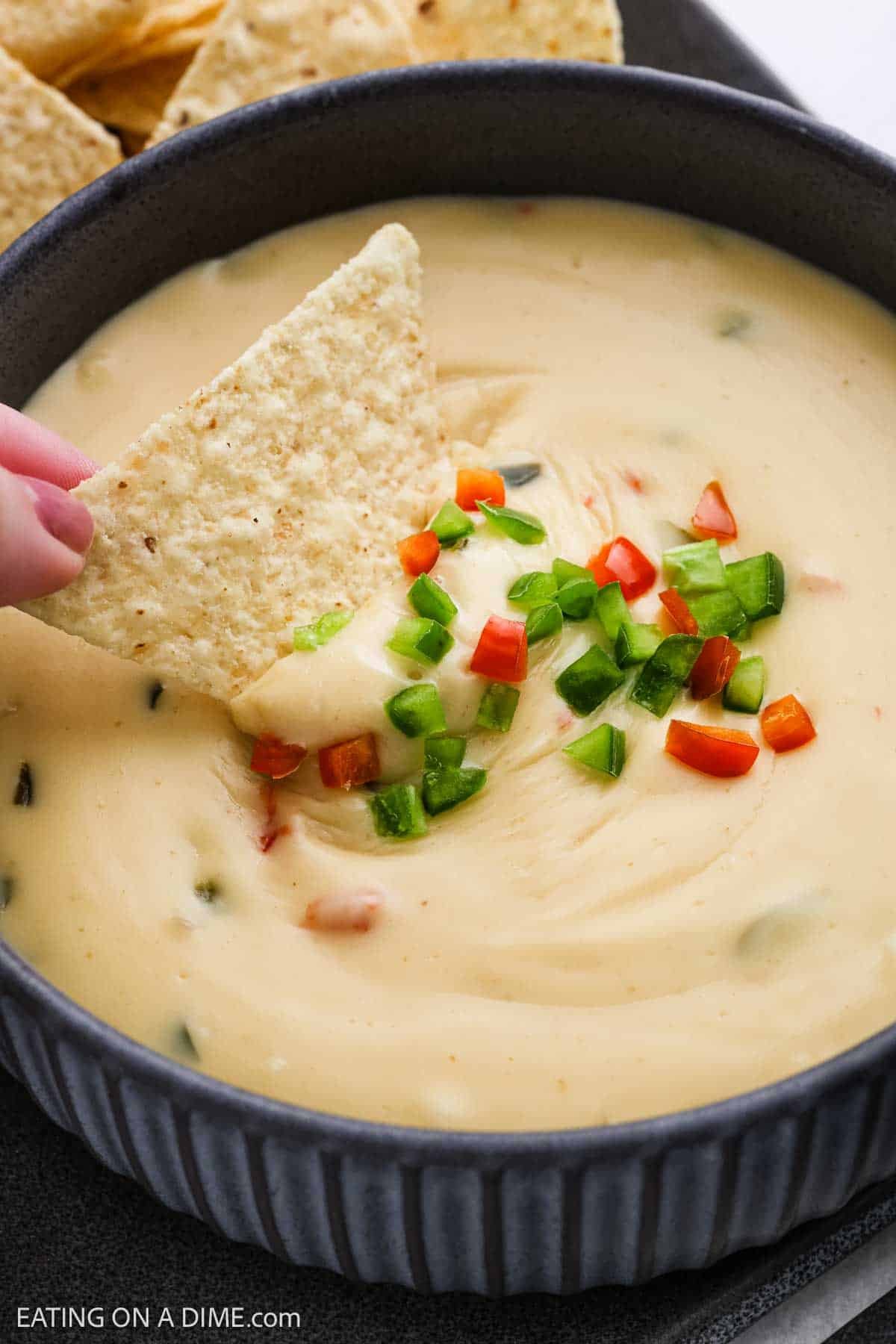 A hand dips a tortilla chip into a bowl of creamy Copycat Chipotle Queso Blanco, topped with chopped green and red peppers. More tortilla chips are visible in the background.