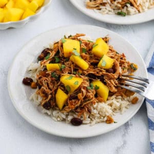 A plate of white rice topped with Crockpot Mango Salsa Chicken, black beans, corn, and mango chunks, garnished with fresh parsley. A fork rests on the plate, and a bowl of diced mango is in the background.