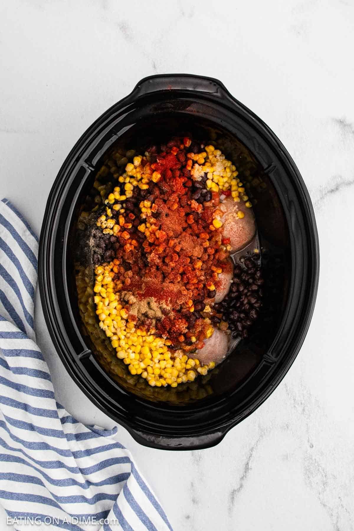 A Crockpot Mango Salsa Chicken recipe awaits as the slow cooker brims with raw chicken breasts, corn, black beans, and spices. A blue and white striped towel sits beside the cooker on a white surface.