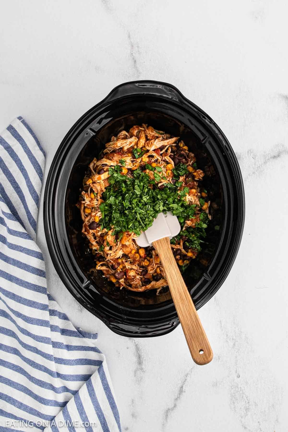 A slow cooker filled with Crockpot Mango Salsa Chicken, shredded with vegetables and topped with chopped fresh herbs. A wooden-handled spatula rests inside. A blue and white striped towel is beside the cooker on a light marble surface.