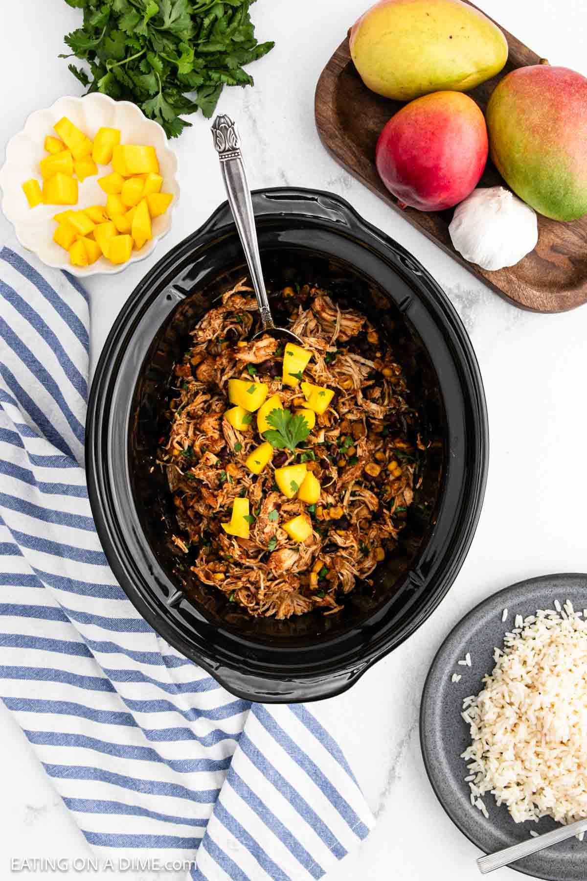 Overhead view of Crockpot Mango Salsa Chicken in a black slow cooker, featuring shredded chicken with mango pieces. Nearby are a plate of rice, a bowl of diced mango, whole mangoes, garlic, cilantro, and a striped blue towel.